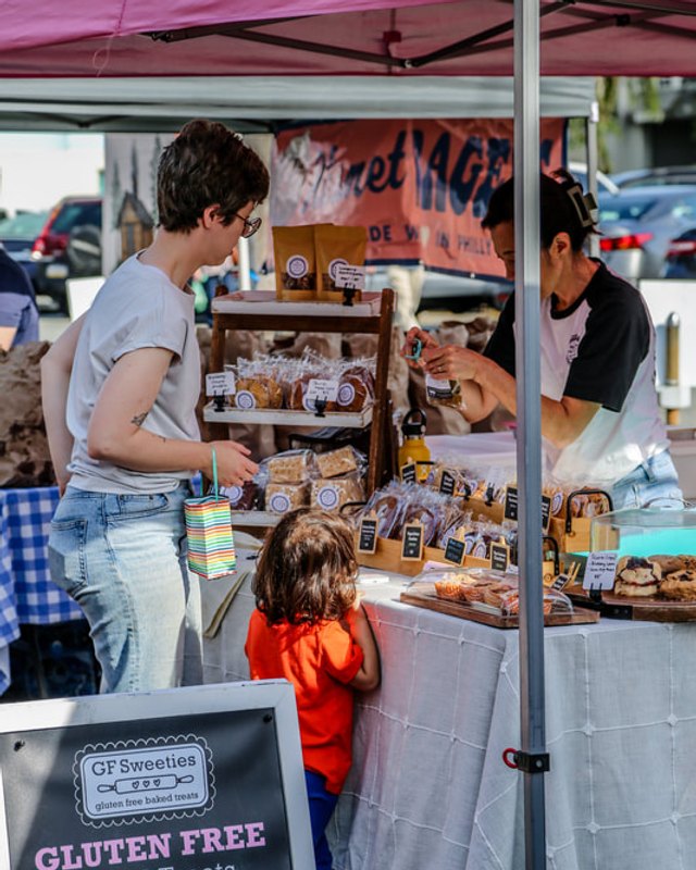 Shoppers at a market booth with baked goods and a gluten-free sign under a pink canopy.