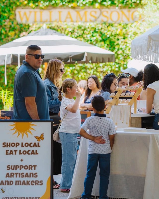 Shoppers browsing market booths under white umbrellas at a 714 Market event with a Shop Local sign in front.