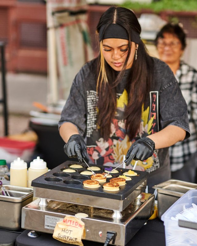 A vendor in black gloves cooking small batter cakes on a griddle at a food booth during an outdoor event