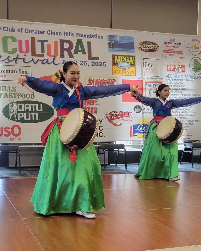 Two women in green and blue hanbok drumming and dancing in front of a Multicultural Festival banner.