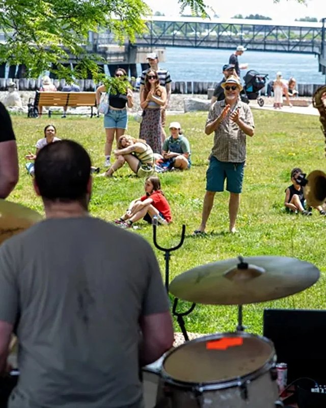 Festival crowd relaxing on a grassy lawn by the water, with a drummer's kit in the foreground at the Resurgam festival in Portland.