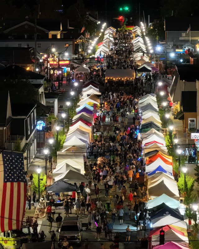 Night aerial view of a dense crowd walking between rows of lit festival tents on a town street