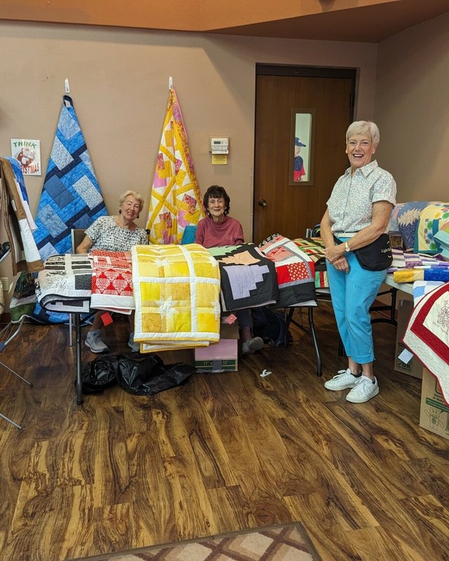 Quilt and craft vendors standing behind a table of handmade quilts at an indoor church craft fair.
