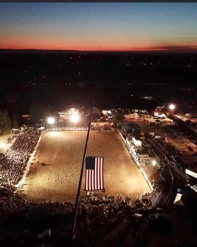 Night view of a packed rodeo arena with crowd stands and an American flag lit up at the Crooked 8 venue.