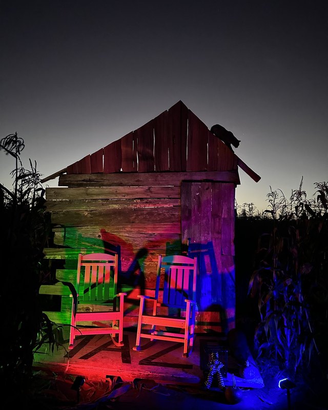 A wooden shed lit with red, green and blue light beside the corn maze at night, with two chairs out front