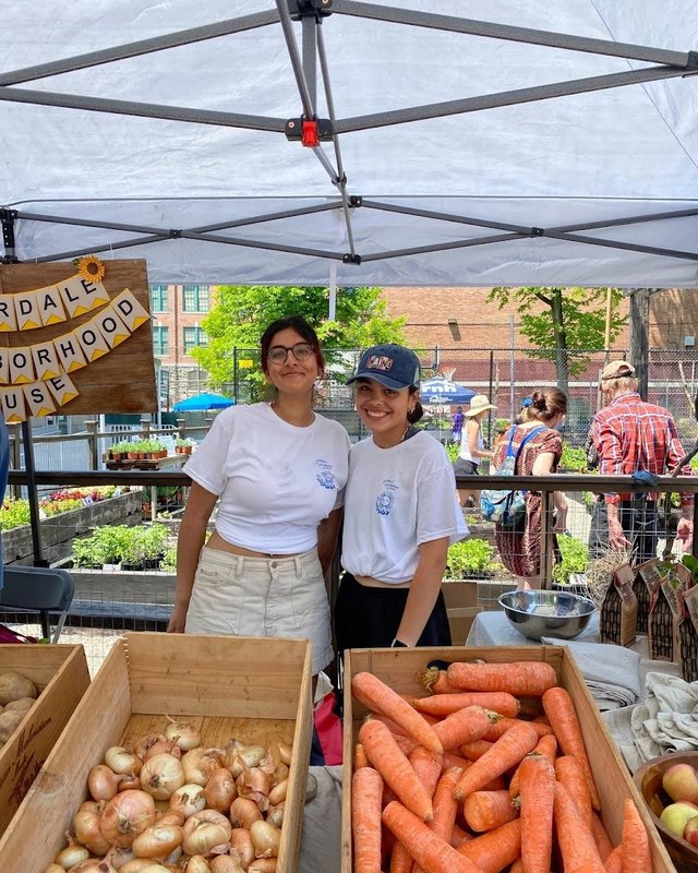 Two volunteers stand at a Riverdale Neighborhood House market booth beside crates of onions and carrots.
