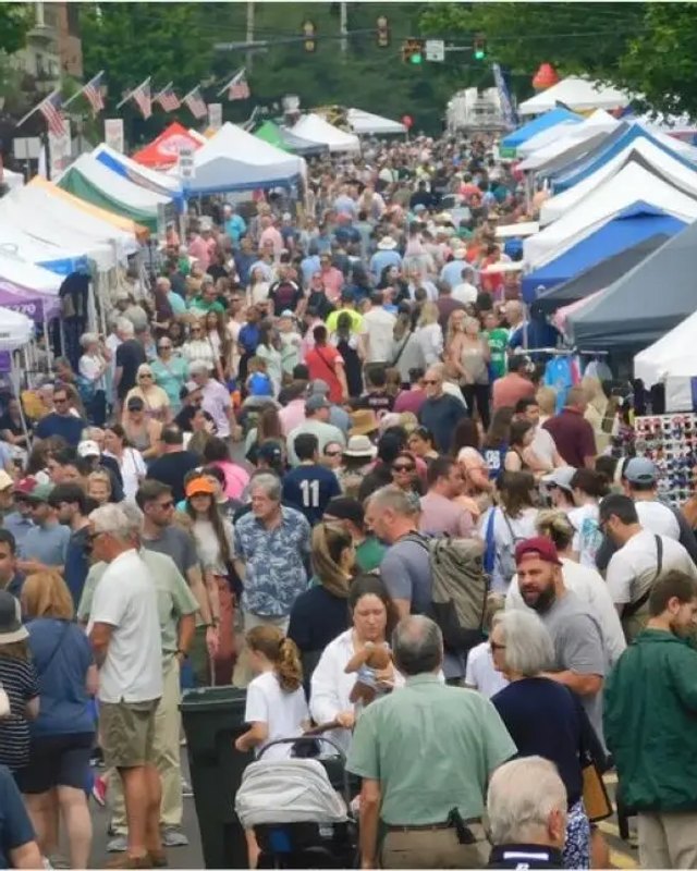 Crowds of shoppers walking between white and colored booth tents at a street fair in Newtown.