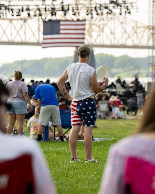 A man in flag-print shorts holds a frisbee and faces a crowd on the lawn, with an American flag hanging over the stage at Waterfront Park.