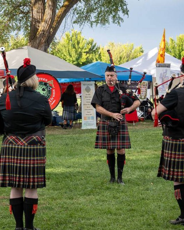 Kilted pipe band playing among market tents and booths on a grassy lawn at the festival.