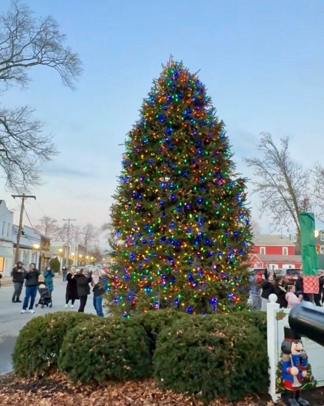 Large illuminated Christmas tree on a town street with people walking nearby.