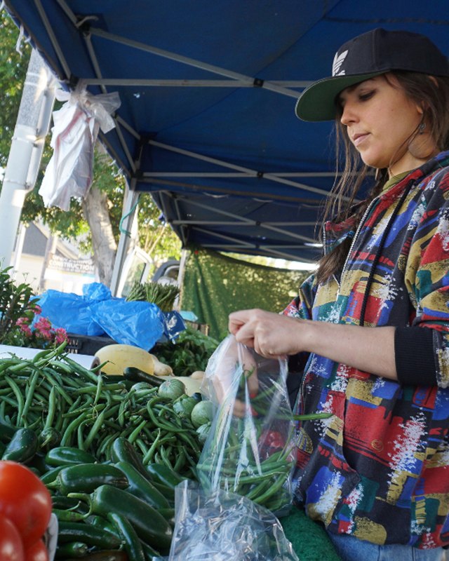 Shoppers at a produce stand under a blue canopy, with tomatoes and green beans on display at the market.