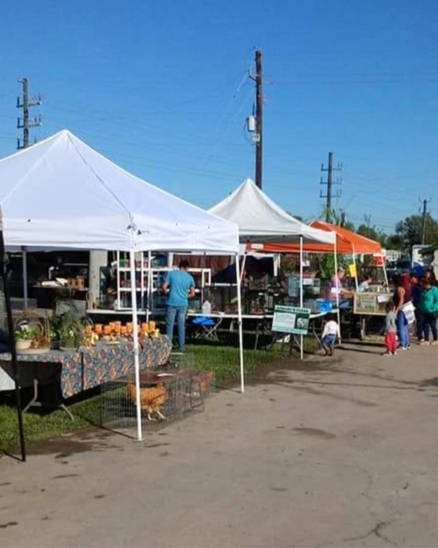 Market tents and shoppers along a dirt lane at an outdoor trade day with produce tables.