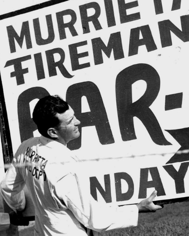 Black-and-white vintage photo of a Murrieta Fireman's BAR-B-Q sign with people standing beside it.