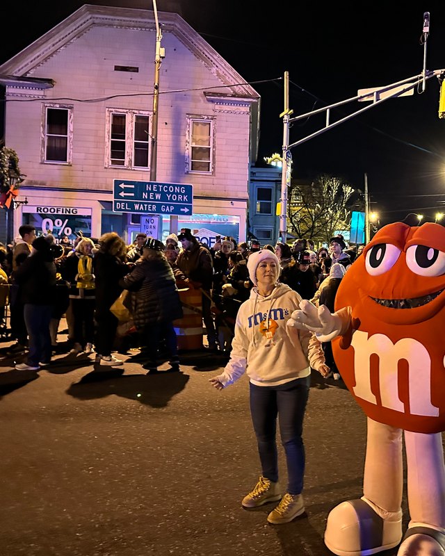 A woman in a cream sweatshirt poses beside a large M&M mascot, with a crowd on a town street at night during the NYE celebration.