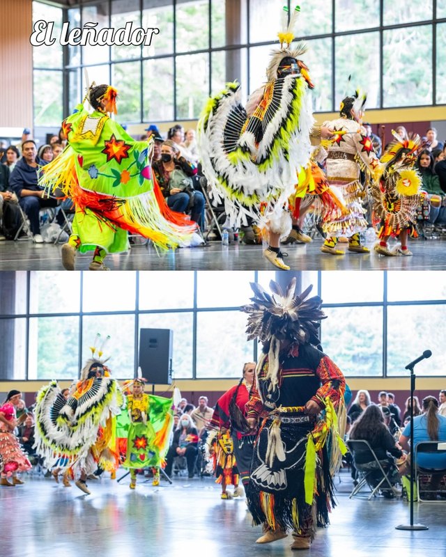 Dancers in colorful regalia and feathered headdresses performing before a crowd in an indoor hall