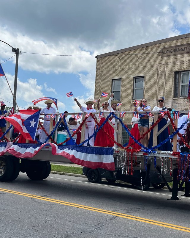 People on a Puerto Rican flag-decorated parade float waving small flags along a Lorain street, with a brick building behind.