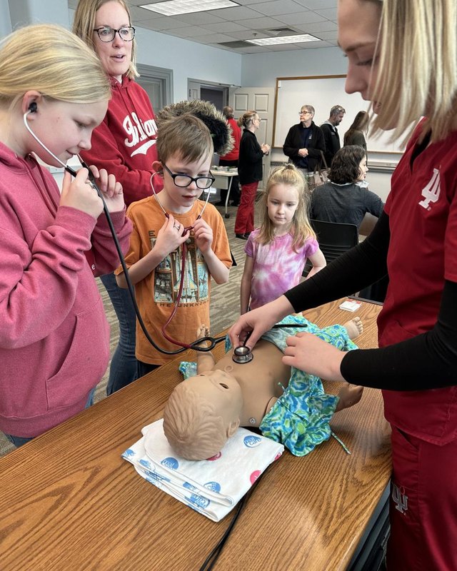 Children and adults at a hands-on medical simulation activity with a patient mannequin during a science event.