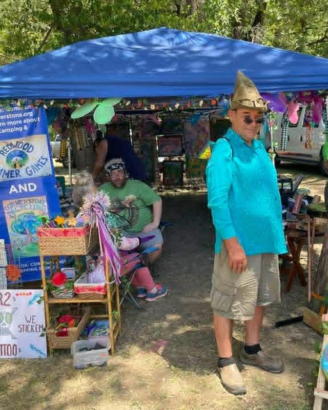 A craft booth under a blue tent at a Fairy Tale Festival, with a costumed person standing beside it