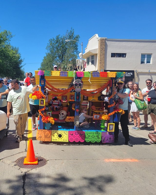 Street crowd watching a decorated festival cart with colorful papel picado and a large white sculpture on a sunny city street.