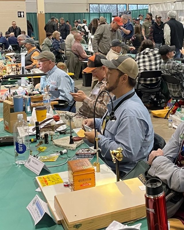 Seated fly tiers and visitors working at tables covered with fly-tying materials inside the Western Idaho Fly Fishing Expo hall.