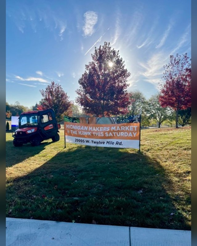 Michigan Makers Market at The Hawk sign on a lawn under a sunny sky, with a utility vehicle and trees nearby.