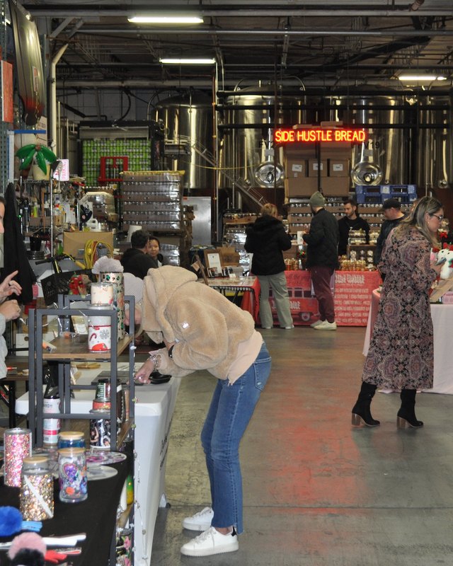 Shoppers browsing crafts and holiday goods at vendor tables inside a warehouse-style market hall