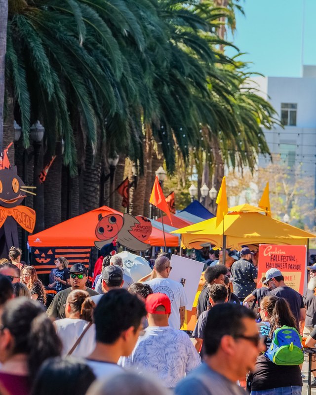 Crowd browsing orange and yellow vendor tents along a palm-lined street at the Anaheim Fall Festival.