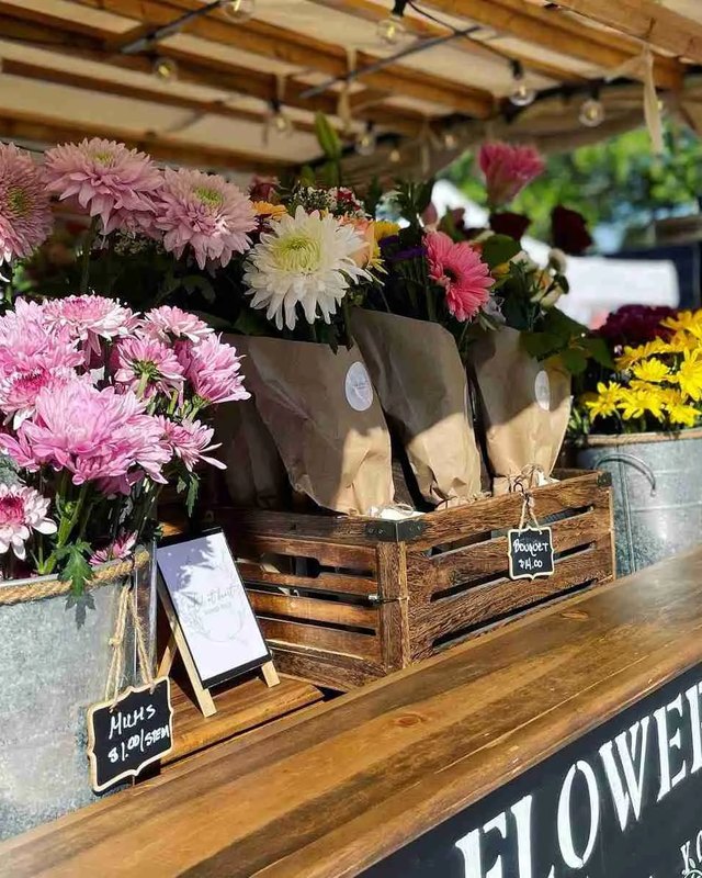 Flower stall with pink and white bouquets in crates at a market, with a chalkboard Flower Bar sign.