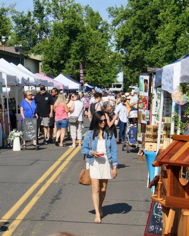 Shoppers walking past white vendor tents and wooden booths along a tree-lined street at the Gresham festival