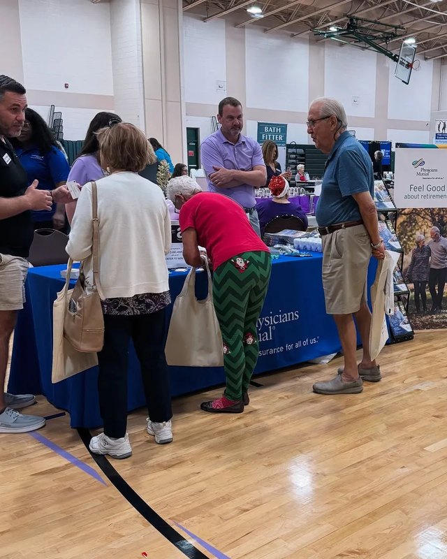 Shoppers and seniors browsing exhibitor booths inside a bright indoor expo hall with blue-draped tables.