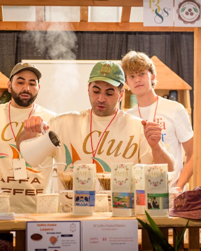 Three coffee vendors at a booth with bags of coffee, one pouring hot water from a kettle as steam rises, at the San Francisco Coffee Festival.