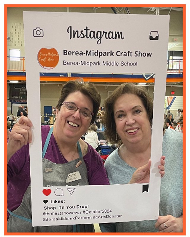 Two smiling women holding a large Instagram-style photo frame at the Berea-Midpark Craft Show, with a hall in the background.