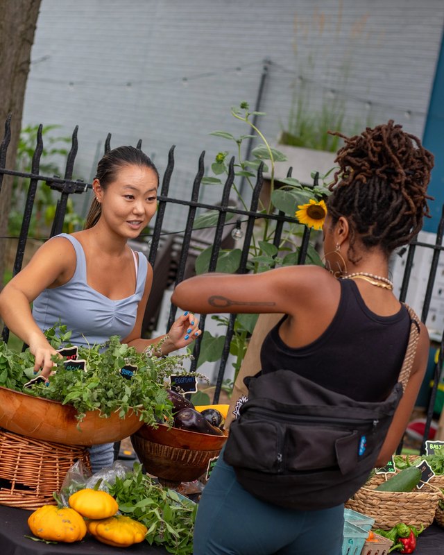 A customer and a vendor chat at a produce stand with baskets of peppers, squash and greens at an outdoor market.