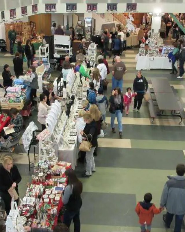 Shoppers browsing craft booths inside an indoor school gym during a holiday bazaar.