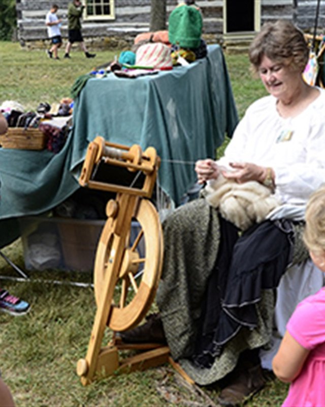 Spinner demonstrating wool spinning at a fall festival booth while children watch under shade trees.