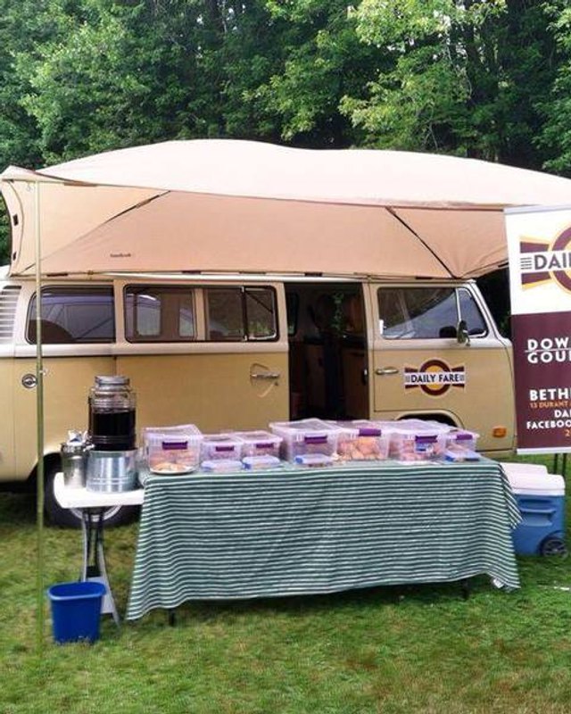 Vintage VW van food truck with a Daily Fare banner and a table of prepared food containers on the grass.
