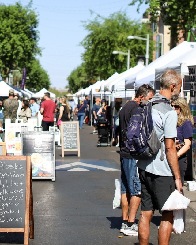 Shoppers walking past white canopy booths along a tree-lined street at a market