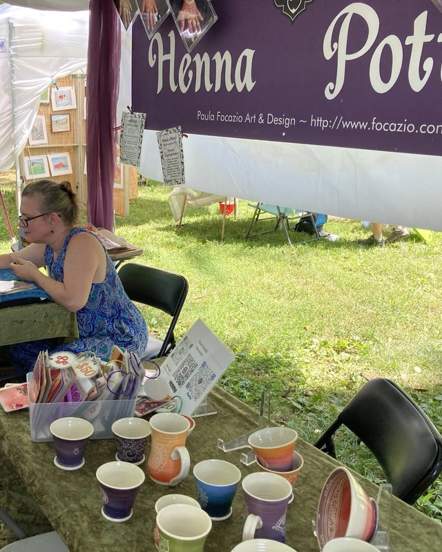 Henna Potter booth at an outdoor arts festival, with ceramic cups displayed on a green table under a white tent.