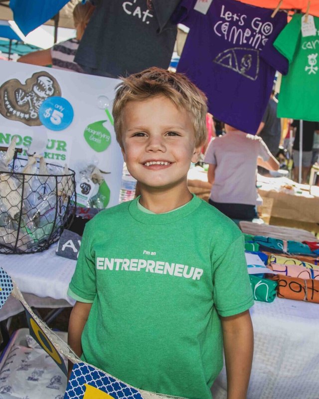 A smiling boy in a green Entrepreneur shirt stands at a market booth covered with kids' t-shirts and goods.
