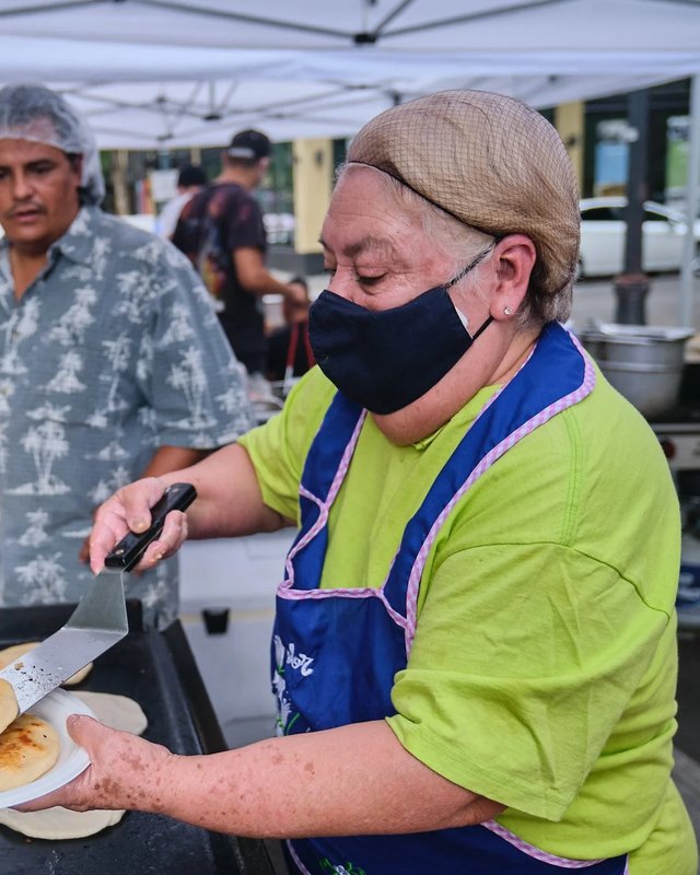 A food vendor in a lime-green shirt and blue apron cooks flatbreads on a griddle at a market stall while people browse nearby.
