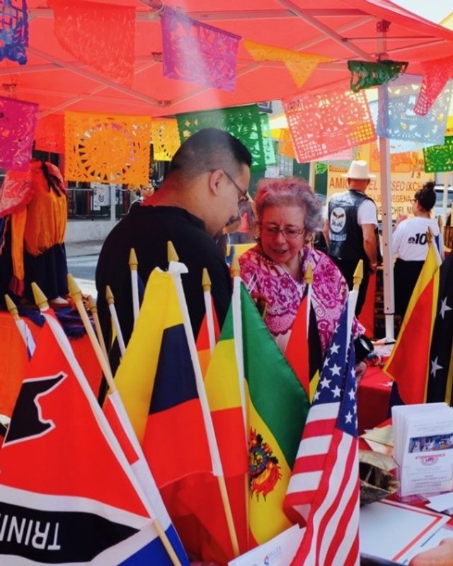 Vendors and visitors at a colorful festival booth with flags and papel picado at La Feria del Barrio