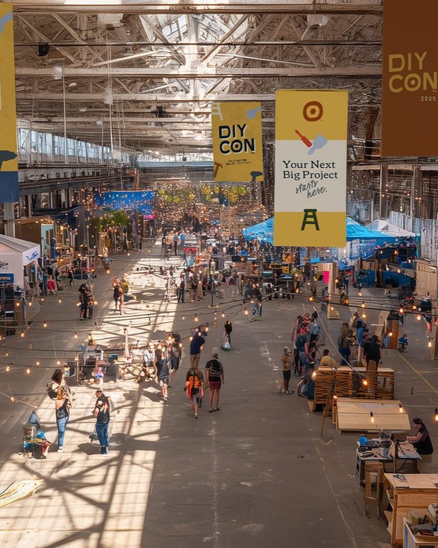 Wide view of the DIY CON hall with rows of wooden booths and shoppers walking under hanging yellow banners.