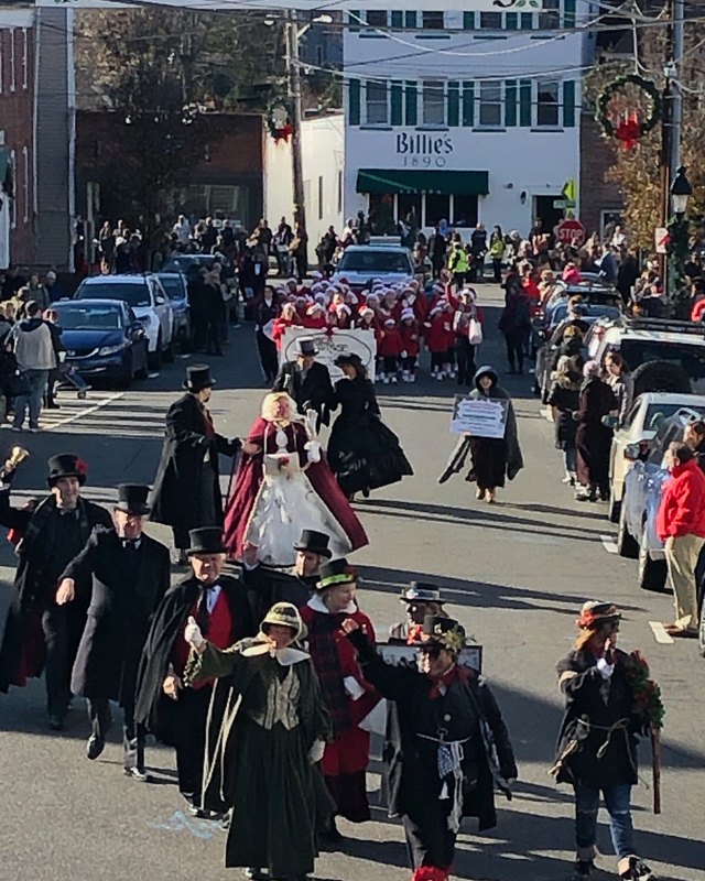 Costumed Dickens characters and crowds marching down a street in a holiday parade in Port Jefferson.