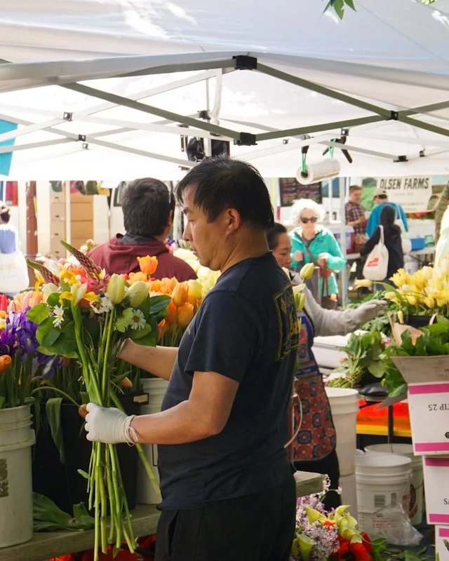 Vendors and shoppers at flower and produce stalls under white tents at a market