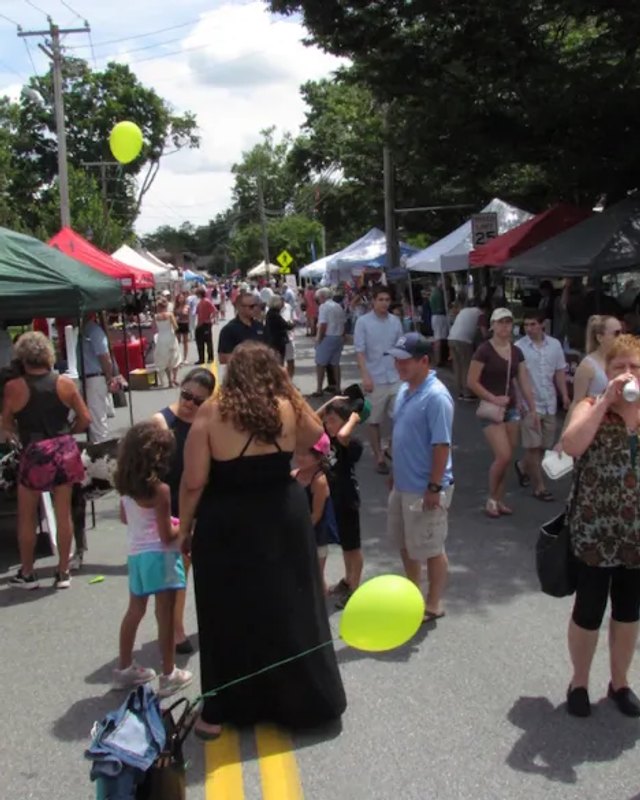 Crowd of shoppers walking past canopy tents, some white and some red, at an outdoor street fair in Wilton; a yellow balloon floats near a woman in a black dress.