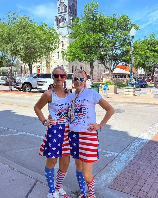 Two women in red, white and blue costumes posing in front of the Granbury courthouse clock tower on the square.
