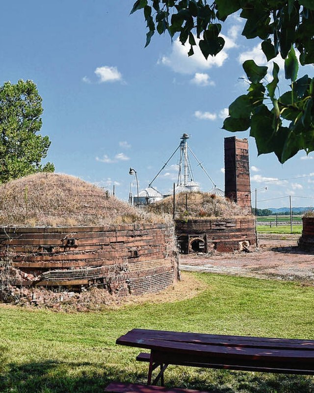 Beehive-shaped brick kilns and a smokestack at the Medora Brick Plant grounds under blue sky.