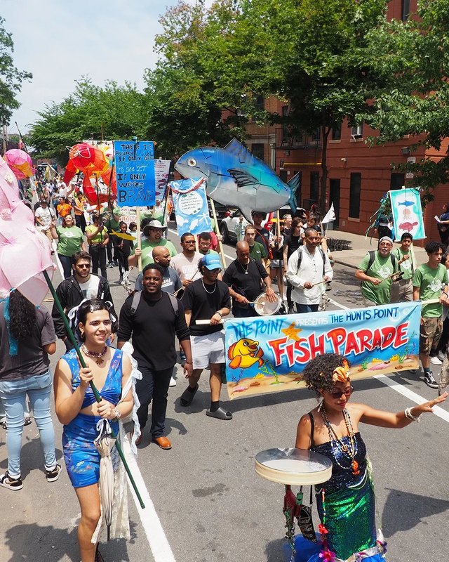 Costumed Fish Parade marchers with a fish float and a Fish Parade banner on a city street.
