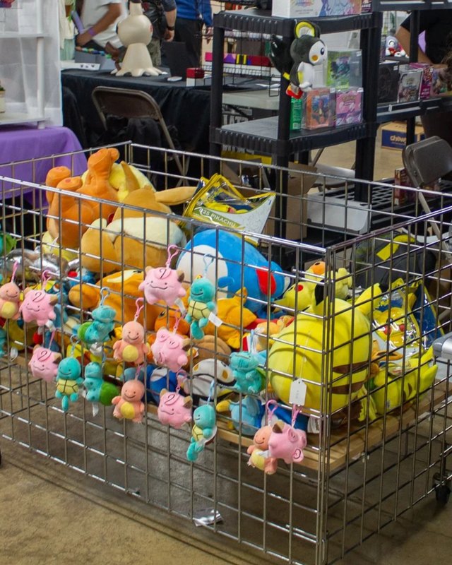 Plush toys piled in a wire cart beside a merchandise vendor table at a PokeFair event.