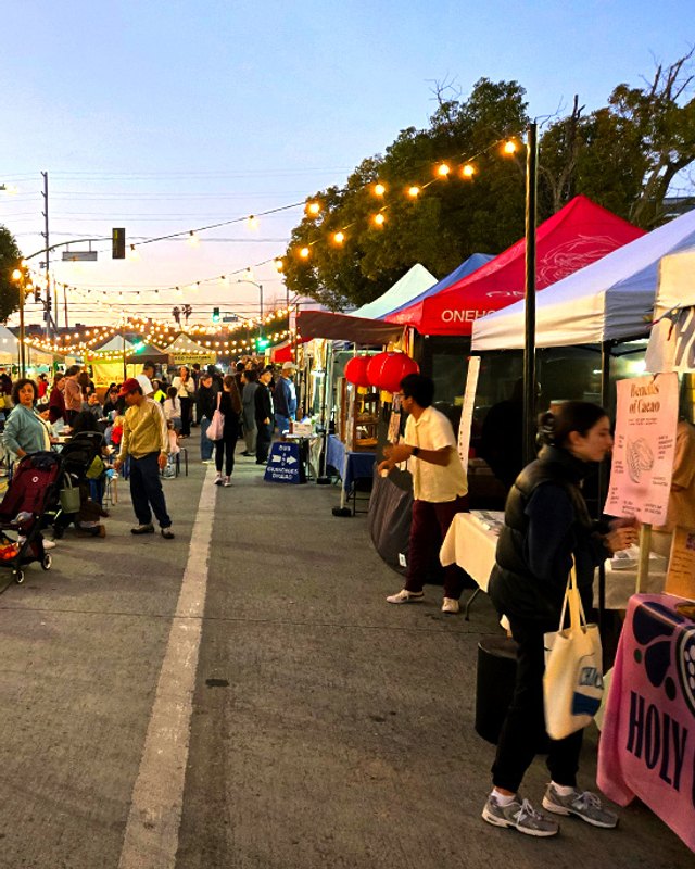 Evening crowd browsing market tents and stalls on a string-lit street at the Culver City Farmers Market.